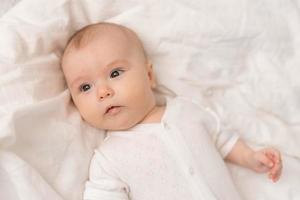 portrait of a cute baby in a white bodysuit on a bed at home with white linens. Newborn baby at home, happy motherhood. High quality photo