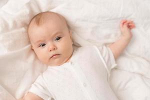 portrait of a cute baby in a white bodysuit on a bed at home with white linens. Newborn baby at home, happy motherhood. High quality photo