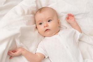 portrait of a cute baby in a white bodysuit on a bed at home with white linens. Newborn baby at home, happy motherhood. High quality photo