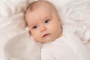 portrait of a cute baby in a white bodysuit on a bed at home with white linens. Newborn baby at home, happy motherhood. High quality photo