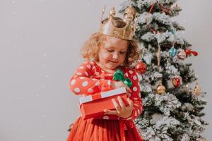 little curly girl in a red carnival dress with a Santa print and a crown is playing with a toy deer next to the Christmas tree. lifestyle, children's emotions. space for text. High quality photo