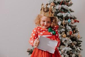 little curly girl in a red carnival dress with a Santa print and a crown is playing with a toy deer next to the Christmas tree. lifestyle, children's emotions. space for text. High quality photo