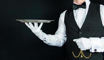 retrato de camarero o mayordomo con chaleco negro y guantes blancos sosteniendo una bandeja de plata sobre fondo negro. concepto de servicio elegante y hospitalidad.