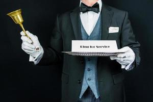 Portrait of Butler or Concierge in Dark Formal Attire and White Gloves Holding Gold Bell and Sign on Silver Tray. Ring for Service. Concept of Professional Courtesy. photo