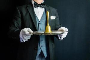 retrato de mayordomo con traje formal oscuro y guantes blancos sosteniendo una campana de oro en una bandeja de plata. concepto de industria de servicios y hospitalidad profesional.