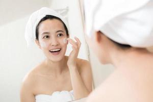 Happy young Asian woman applying face lotions while wearing a towel and touching her face in bathroom photo
