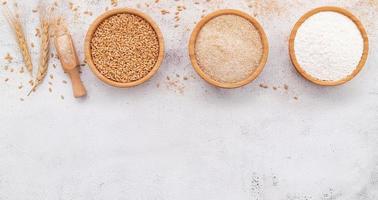 granos de trigo, harina de trigo marrón y harina de trigo blanco en un tazón de madera sobre fondo de hormigón blanco. foto