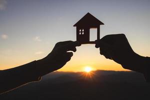 silueta de manos masculinas y femeninas sosteniendo una casa modelo al atardecer concepto de comprar casas, bienes raíces. foto