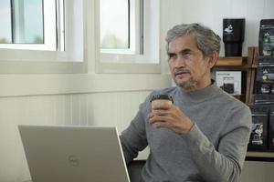 Portrait elderly senior asian man sits near glass window in the morning to work from home and checking his business on his laptop on table seriously, soft and selective focus. photo