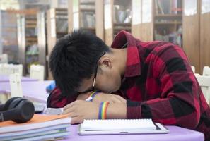 Young Asian boy wears rainbow wristband, sitting in library and taking a nap on table while doing his hard school project work, concept for raising teens and LGBT people daily life activity. photo