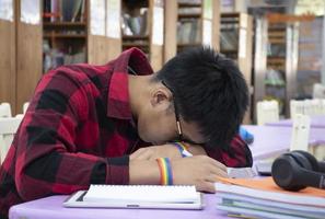 Young Asian boy wears rainbow wristband, sitting in library and taking a nap on table while doing his hard school project work, concept for raising teens and LGBT people daily life activity. photo