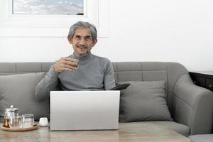 Elderly asian grey hair man in long sleeve shirt sits on sofa and drinking hot tea in living room of his house, concept for happy life of elders and life insurance around the world. photo