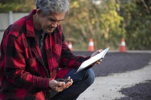 un anciano agricultor asiático inteligente de granos de café con pantalones rojo y negro sentado en el área central del piso de secado de la pila de granos de café y sosteniendo taplet en la mano para verificar la calidad del café, enfoque suave y selectivo. foto