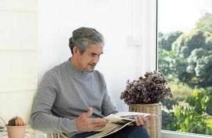 retrata a un anciano asiático sentado cerca de una ventana de vidrio por la mañana para trabajar desde casa y revisando su negocio en su computadora portátil en la mesa con seriedad, enfoque suave y selectivo. foto