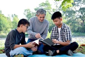 An elderly asian male is spending his spare time explaining the contents of the lesson and happily helping his two grandsons with their homework and school projects in his backyard, soft focus. photo