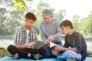 An elderly asian male is spending his spare time explaining the contents of the lesson and happily helping his two grandsons with their homework and school projects in his backyard, soft focus. photo