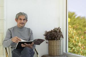 Portrait elderly senior asian man sits near glass window in the morning to work from home and checking his business on his laptop on table seriously, soft and selective focus. photo