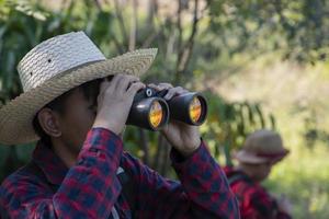 Asian boy is using binoculars to do the birds' watching in tropical forest during summer camp, idea for learning creatures and wildlife animals and insects outside the classroom. photo