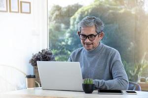 retrata a un anciano asiático sentado cerca de una ventana de vidrio por la mañana para trabajar desde casa y revisando su negocio en su computadora portátil en la mesa con seriedad, enfoque suave y selectivo.