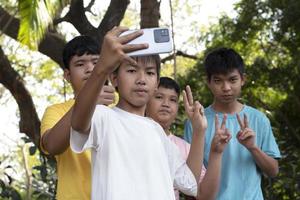 Group of young asian teen boys spending free times in the park rasing their fingers and taking selfie together happily, soft and selective focus on boy in white t-shirt, raising teens concept. photo