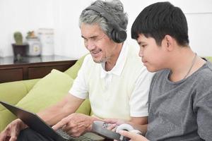 Asian senior grandpa and his son are spending times together at home to connect headphone with his laptop to watch and to play online games, soft and selective focus, raising teens concept. photo