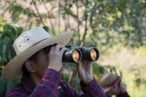 Asian boy is using binoculars to do the birds' watching in tropical forest during summer camp, idea for learning creatures and wildlife animals and insects outside the classroom. photo