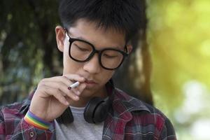 Young asian teen boy in plaid shirt wears rainbow wristband and holds cigarette in hand and smoking, blurred background, concept for bad behavior of young teens all over the world.