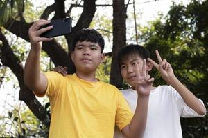 Group of young asian teen boys spending free times in the park rasing their fingers and taking selfie together happily, soft and selective focus on boy in white t-shirt, raising teens concept. photo