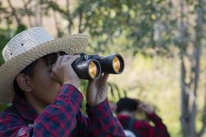 Asian boy is using binoculars to do the birds' watching in tropical forest during summer camp, idea for learning creatures and wildlife animals and insects outside the classroom. photo