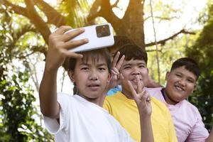 Group of young asian teen boys spending free times in the park rasing their fingers and taking selfie together happily, soft and selective focus on boy in white t-shirt, raising teens concept. photo