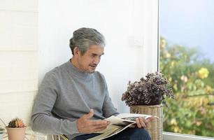 Portrait elderly senior asian man sits near glass window in the morning to work from home and checking his business on his laptop on table seriously, soft and selective focus. photo