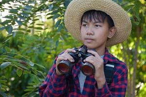 un niño asiático está usando binoculares para observar aves en el bosque tropical durante el campamento de verano, idea para aprender criaturas y animales salvajes e insectos fuera del aula. foto