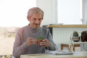 Portrait elderly senior asian man sits near glass window in the morning to work from home and checking his business on his laptop on table seriously, soft and selective focus. photo