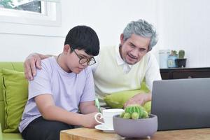 Asian senior grandpa and his son are spending times together at home to connect headphone with his laptop to watch and to play online games, soft and selective focus, raising teens concept. photo