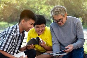 An elderly asian male is spending his spare time explaining the contents of the lesson and happily helping his two grandsons with their homework and school projects in his backyard, soft focus. photo