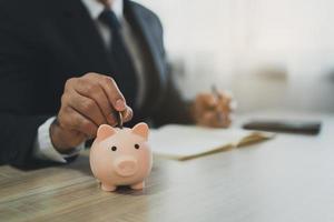 Business man with suit drop coins piggy bank on table, saving money wealth and financial concept, Business, finance, investment, Financial planning. Business financial saving for future concept. photo