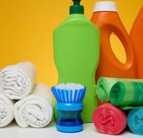 botellas de plástico naranja con productos líquidos y bolsas de basura, cepillos y trapos textiles para lavar suelos sobre una mesa blanca