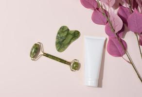 masajeador facial manual de jade y tubos de plástico blanco para cosméticos sobre un fondo beige