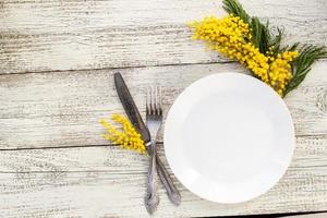 plato de mesa festivo con tenedor y cuchillo y decoración floral de mimosa sobre fondo blanco de madera con espacio para copiar foto