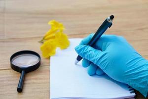 Student  hands in protective gloves hold magnifying glass to inspect flowers, holds pen to write on paper. Concept, science subject, project work. Experiment, education, learning by doing approach. photo