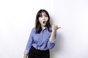 Shocked Asian woman wearing blue shirt pointing at the copy space beside her, isolated by white background photo