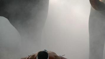 Handaufnahme, junger Mann mit Kopf und beiden Händen, die in verdrehtes Hanfseil gewickelt sind und Kunst des Muay Thai zeigen, verschwommene Elefanten und sich ausbreitender weißer Nebel im Hintergrund