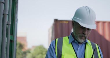 Handheld medium shot, middle-aged Caucasian Business engineer man wearing helmet checking transport container and writing on clipboard at storage port terminal video