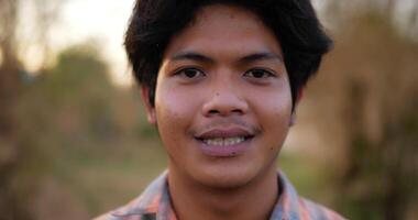 retrato próximo de um jovem fazendeiro feliz asiático olhando para a câmera e sorrindo com um fundo de terras agrícolas. conceito de agricultura agrícola. câmera lenta. video