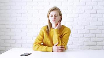 mulher pensativa de suéter amarelo sentada à mesa pensativa, usando smartphone video