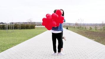 Liebe. Junger Mann umarmt und küsst ein Mädchen im Park mit roten Luftballons video