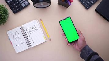 Top view of smart phone with green screen and notepad on table . video