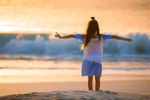 Adorable happy little girl walking on white beach at sunset. photo