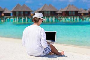 joven trabajando en una laptop en una playa tropical cerca de una villa acuática foto