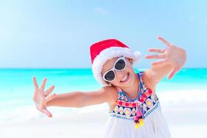 Happy little girl in Santa hat on Christmas beach vacation photo